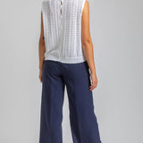 Woman wearing a white sleeveless top with lace details and navy pants on a plain background