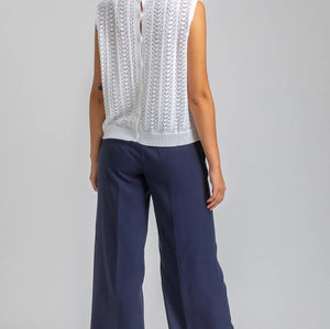 Woman wearing a white sleeveless top with lace details and navy pants on a plain background