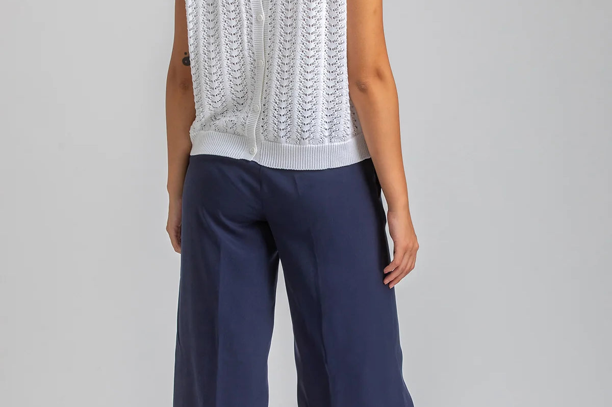 Woman wearing a white sleeveless top with lace details and navy pants on a plain background