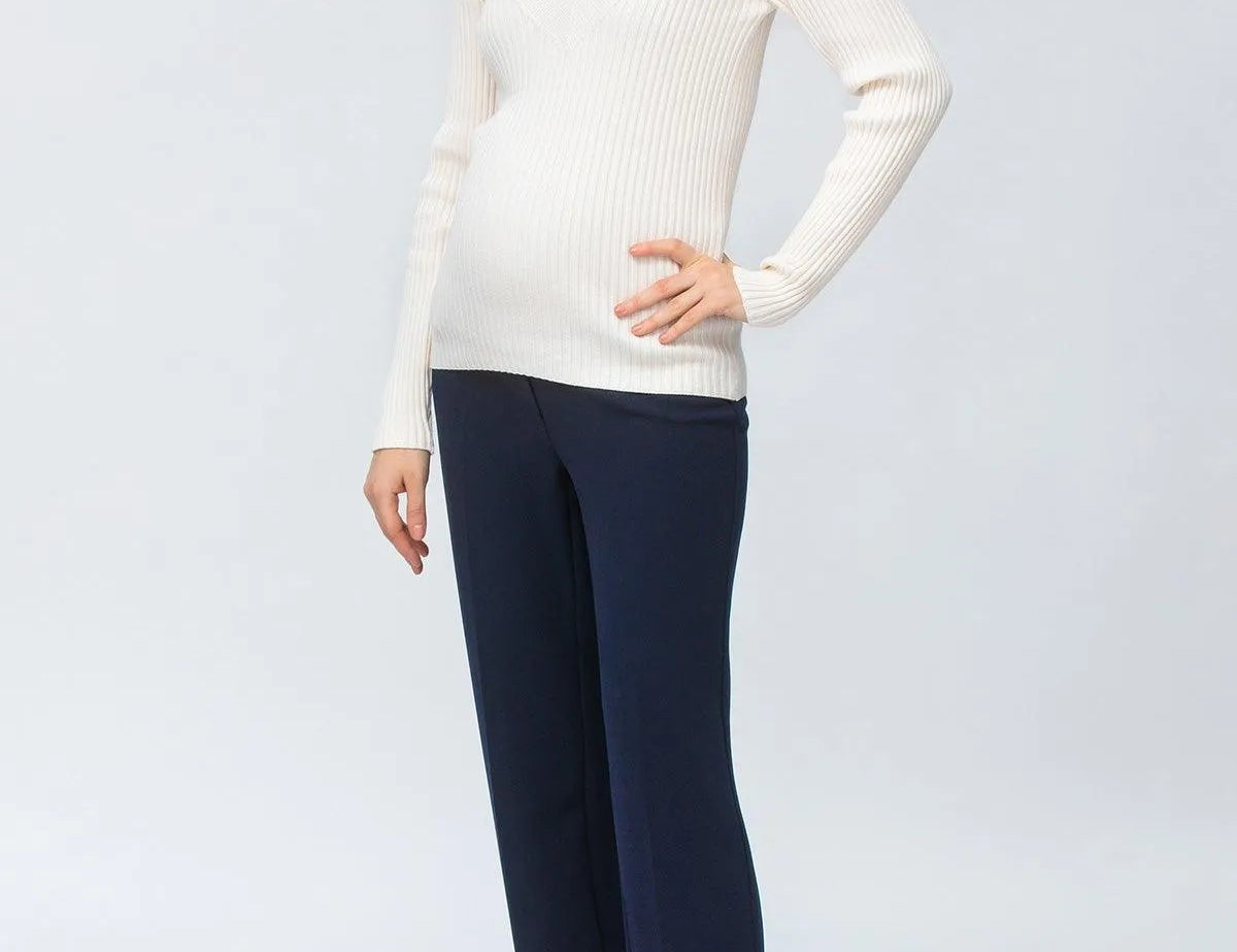 A woman modeling a ribbed white long-sleeve V-neck sweater and navy wide-leg trousers, paired with dark brown pointed-toe heeled boots, against a light gray studio background.