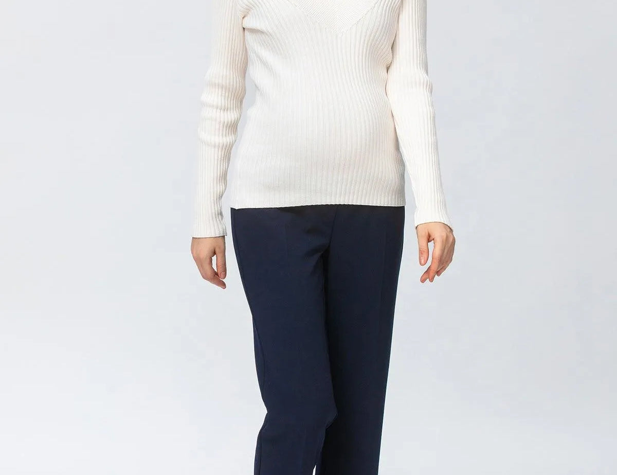 A woman modeling a ribbed white long-sleeve V-neck sweater paired with dark navy trousers and dark brown pointed-toe heels, photographed against a seamless light gray studio background.