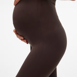 A close-up side view of a pregnant person wearing dark brown maternity leggings, holding the belly, against a plain white background.