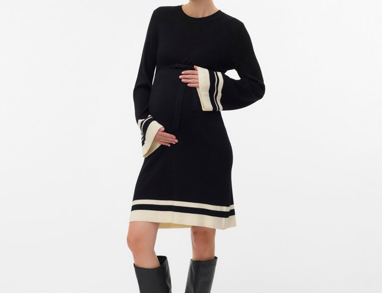 A pregnant model wearing a black long-sleeve knit dress with cream stripe accents at the cuffs and hem, paired with black knee-high boots, against a plain white studio background.