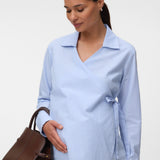 A pregnant woman wearing a light blue and white vertically striped wrap blouse with a collar and side tie, holding a brown leather handbag against a plain light gray studio background.