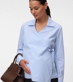 A pregnant woman wearing a light blue and white vertically striped wrap blouse with a collar and side tie, holding a brown leather handbag against a plain light gray studio background.