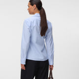 A woman shown from the back wearing a light blue and white vertically striped button-down shirt, paired with black trousers and holding a brown leather handbag against a plain light gray studio background.