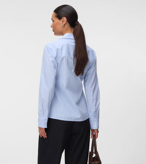 A woman shown from the back wearing a light blue and white vertically striped button-down shirt, paired with black trousers and holding a brown leather handbag against a plain light gray studio background.