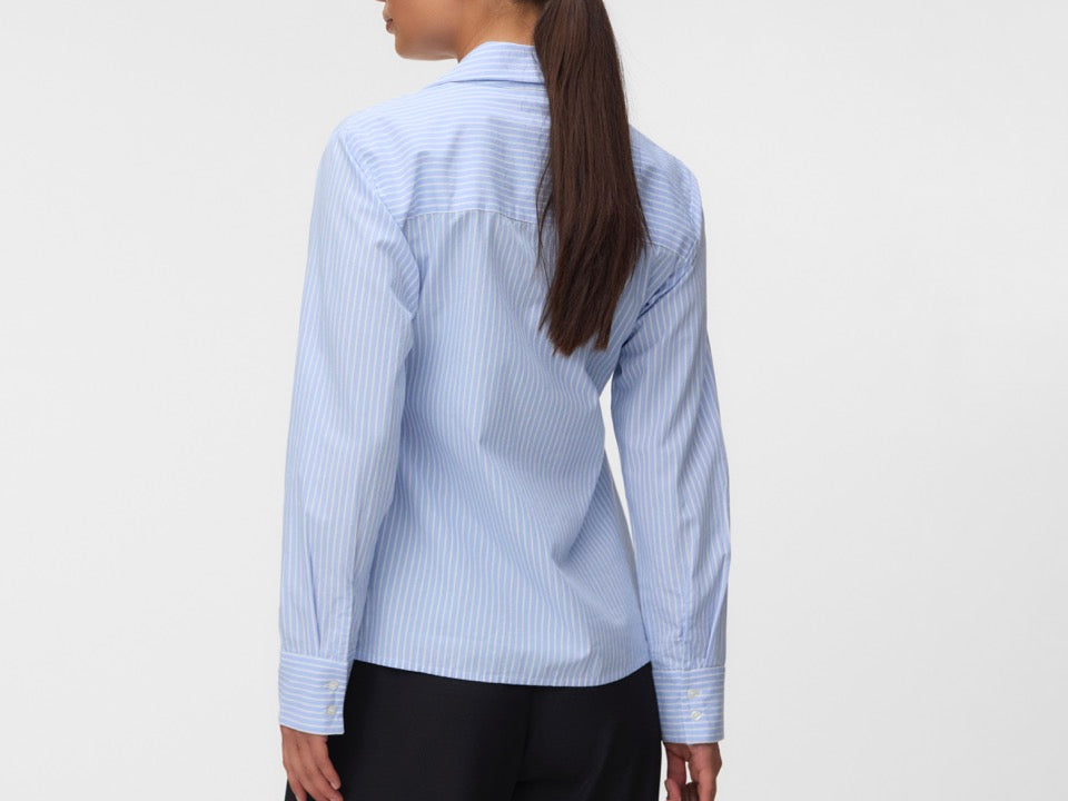 A woman shown from the back wearing a light blue and white vertically striped button-down shirt, paired with black trousers and holding a brown leather handbag against a plain light gray studio background.