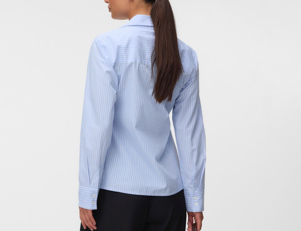 A woman shown from the back wearing a light blue and white vertically striped button-down shirt, paired with black trousers and holding a brown leather handbag against a plain light gray studio background.