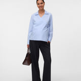 A woman modeling a light blue long-sleeve collared shirt with dark wide-leg trousers, holding a brown handbag against a plain light gray studio background.