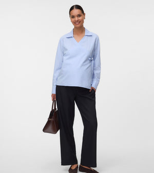 A woman modeling a light blue long-sleeve collared shirt with dark wide-leg trousers, holding a brown handbag against a plain light gray studio background.