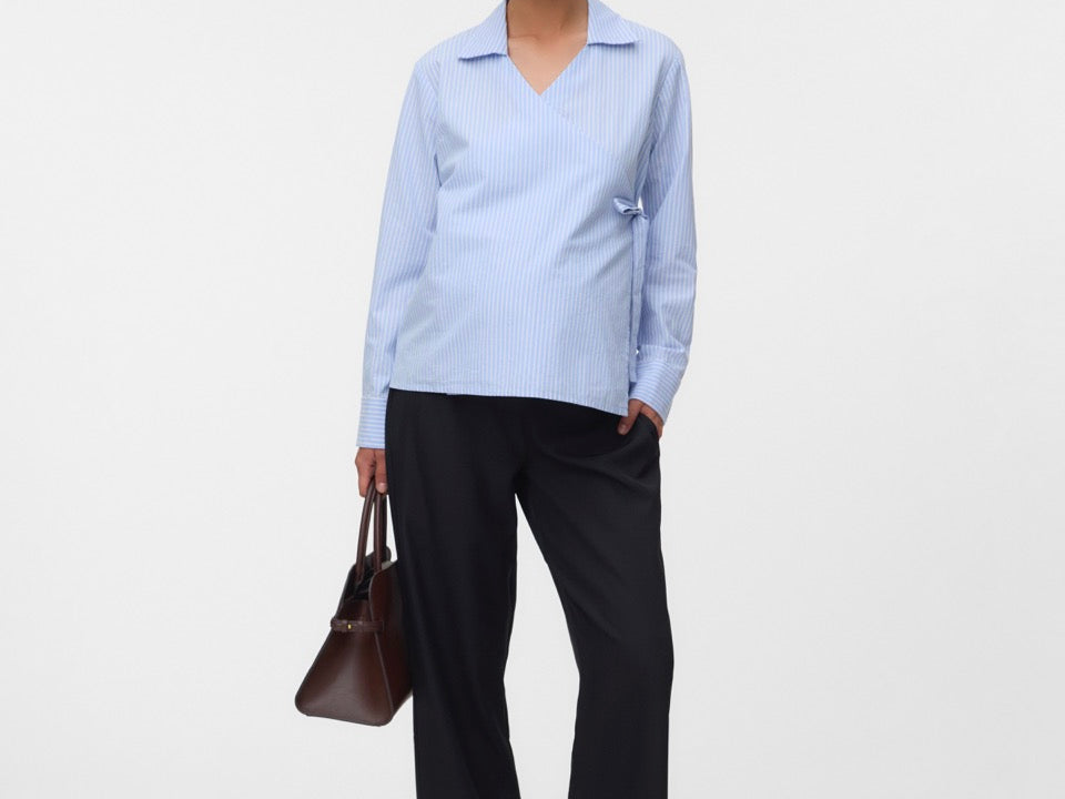 A woman modeling a light blue long-sleeve collared shirt with dark wide-leg trousers, holding a brown handbag against a plain light gray studio background.