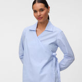 A woman modeling a light blue and white vertically striped wrap blouse with a collar and side tie, paired with black trousers, against a plain light gray studio background.
