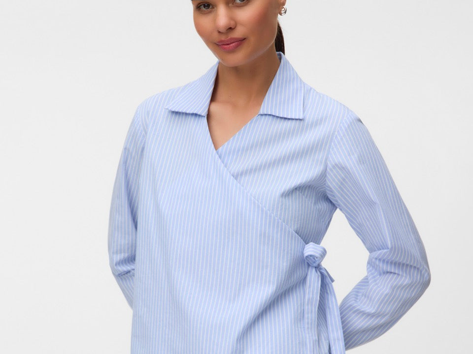A woman modeling a light blue and white vertically striped wrap blouse with a collar and side tie, paired with black trousers, against a plain light gray studio background.