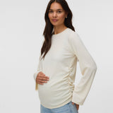 A pregnant woman wearing a cream-colored long-sleeve maternity top with ruching on the sides, paired with light blue jeans, standing against a plain light gray background.