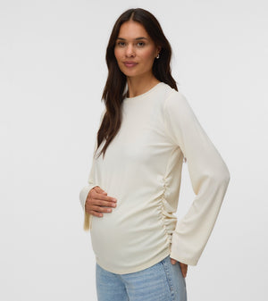 A pregnant woman wearing a cream-colored long-sleeve maternity top with ruching on the sides, paired with light blue jeans, standing against a plain light gray background.