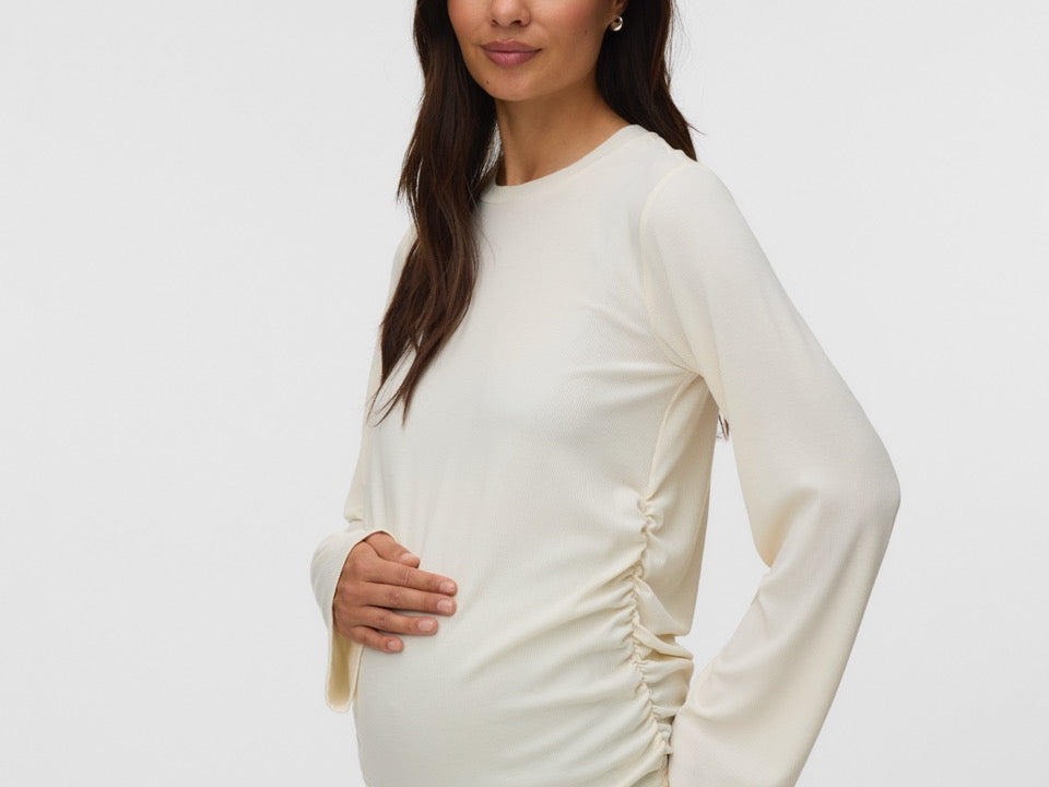 A pregnant woman wearing a cream-colored long-sleeve maternity top with ruching on the sides, paired with light blue jeans, standing against a plain light gray background.