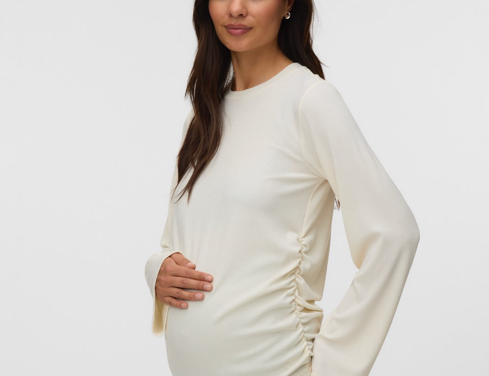 A pregnant woman wearing a cream-colored long-sleeve maternity top with ruching on the sides, paired with light blue jeans, standing against a plain light gray background.