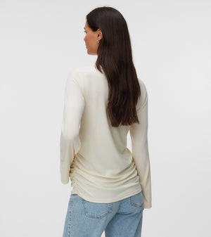 A woman with long dark brown hair is shown from the back wearing a cream-colored long-sleeve knit top and light blue jeans against a plain light gray studio background.