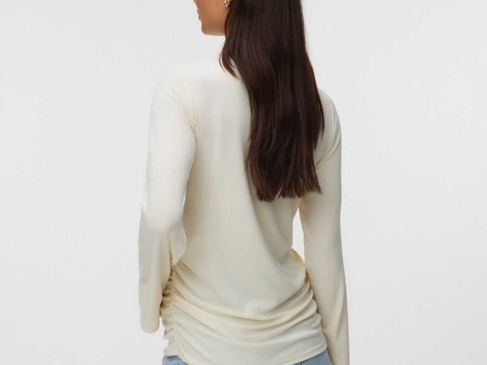 A woman with long dark brown hair is shown from the back wearing a cream-colored long-sleeve knit top and light blue jeans against a plain light gray studio background.
