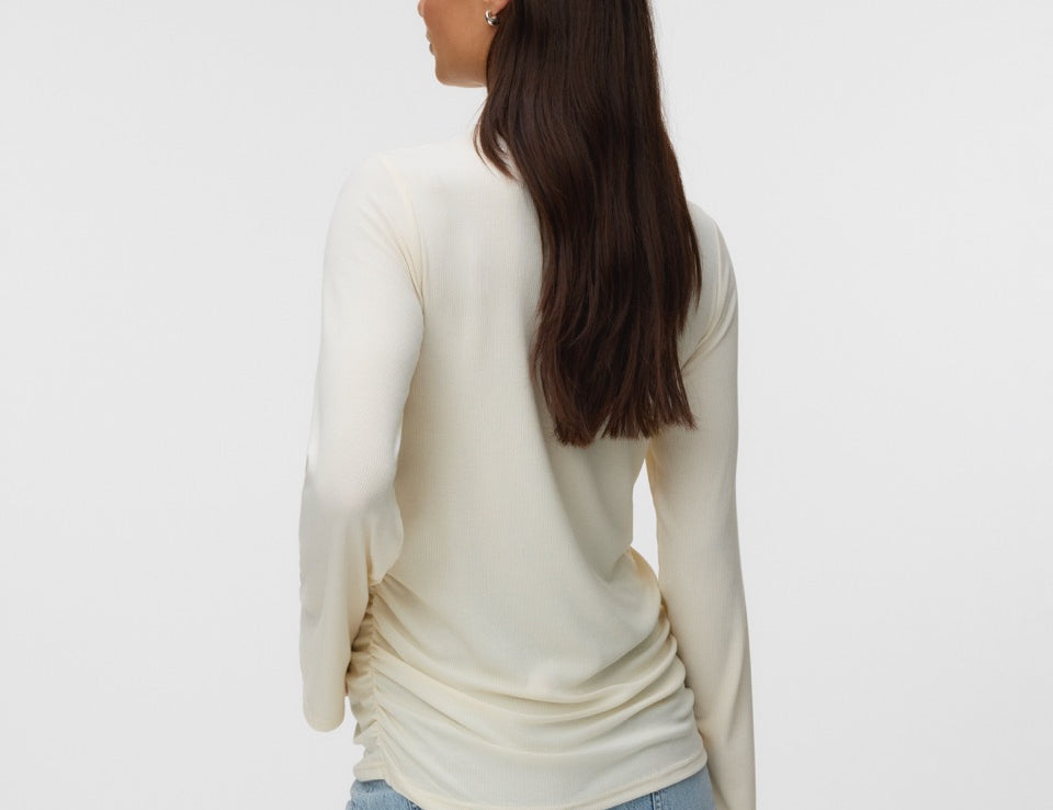 A woman with long dark brown hair is shown from the back wearing a cream-colored long-sleeve knit top and light blue jeans against a plain light gray studio background.
