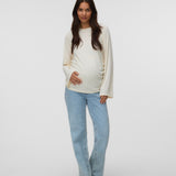 A pregnant woman stands against a light gray studio background wearing a cream-colored long-sleeve maternity top and light blue jeans.