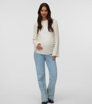 A pregnant woman stands against a light gray studio background wearing a cream-colored long-sleeve maternity top and light blue jeans.