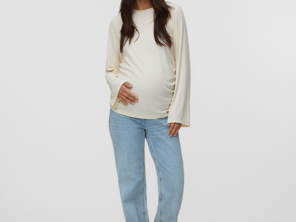 A pregnant woman stands against a light gray studio background wearing a cream-colored long-sleeve maternity top and light blue jeans.