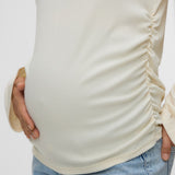 A close-up of a pregnant model wearing a cream-colored, ruched maternity top and light blue jeans, holding her baby bump against a plain light gray background.