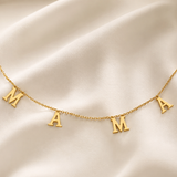 A delicate gold chain necklace featuring three capital letters spelling 'MAMA' spaced evenly along the chain, displayed on a draped cream satin fabric background.