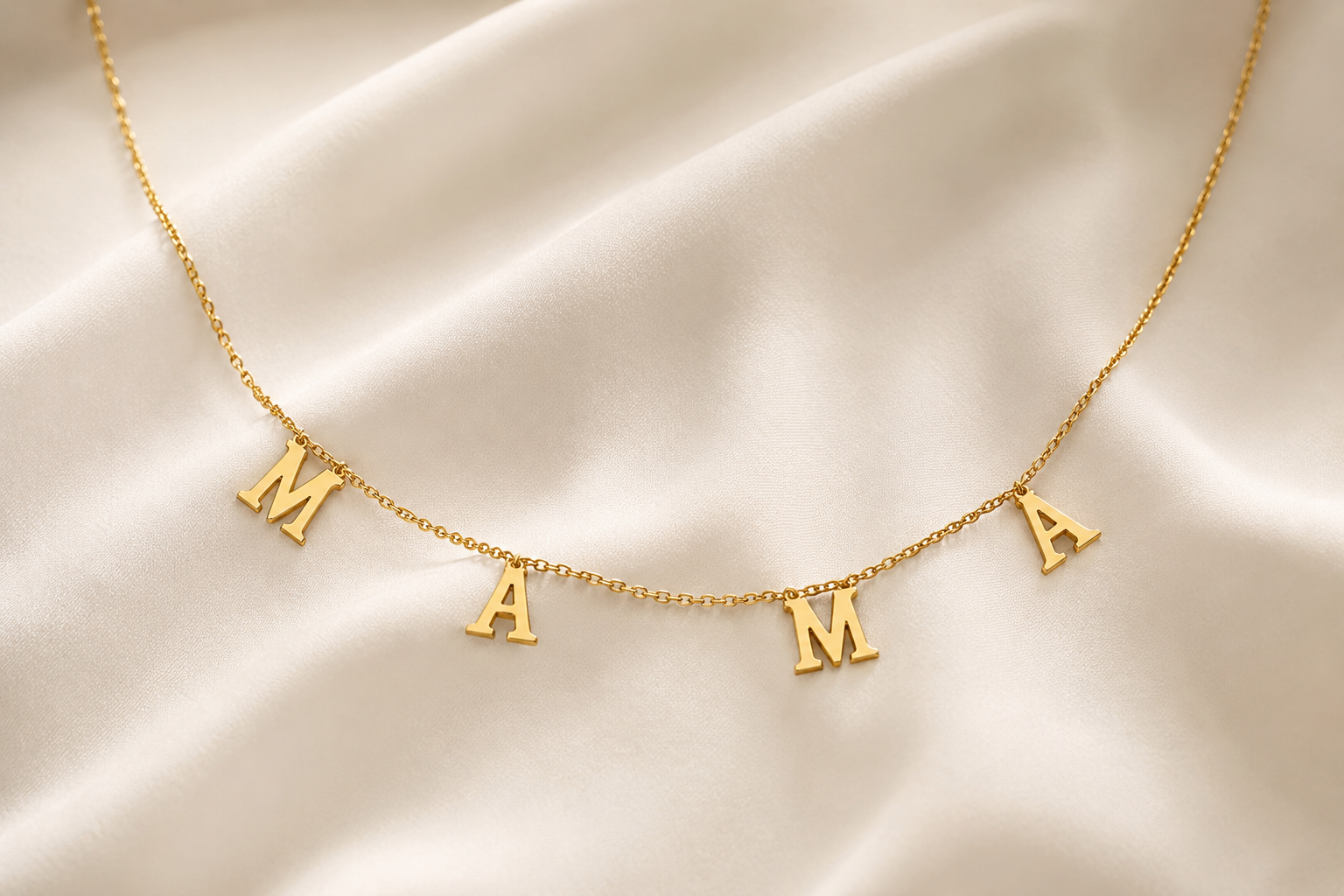 A delicate gold chain necklace featuring three capital letters spelling 'MAMA' spaced evenly along the chain, displayed on a draped cream satin fabric background.