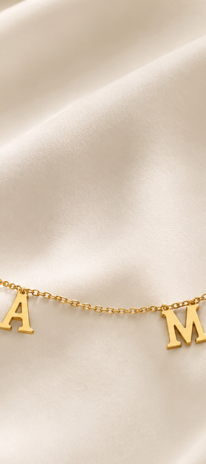 A delicate gold chain necklace featuring three capital letters spelling 'MAMA' spaced evenly along the chain, displayed on a draped cream satin fabric background.