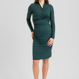 A woman modeling a dark green long-sleeve wrap-style dress with ruching, paired with dark brown pointed-toe heels, against a light gray studio background.