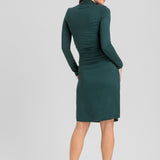 A woman modeling a dark green long-sleeve mock-neck dress from the back, standing against a light gray studio background.