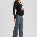 A pregnant woman posing in a black long-sleeve ruched top and gray wide-leg trousers with black high heels, set against a solid light gray studio background.