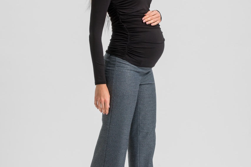 A pregnant woman posing in a black long-sleeve ruched top and gray wide-leg trousers with black high heels, set against a solid light gray studio background.