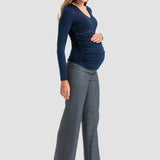 A pregnant woman posing in a studio setting, wearing a navy blue long-sleeve ruched top, gray wide-leg trousers, and black high heels against a light gray background.