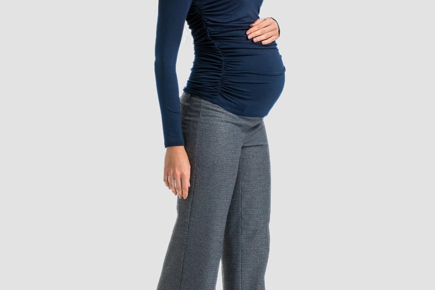 A pregnant woman posing in a studio setting, wearing a navy blue long-sleeve ruched top, gray wide-leg trousers, and black high heels against a light gray background.