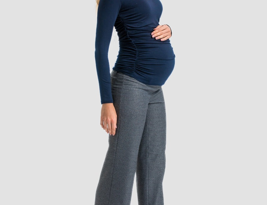 A pregnant woman posing in a studio setting, wearing a navy blue long-sleeve ruched top, gray wide-leg trousers, and black high heels against a light gray background.