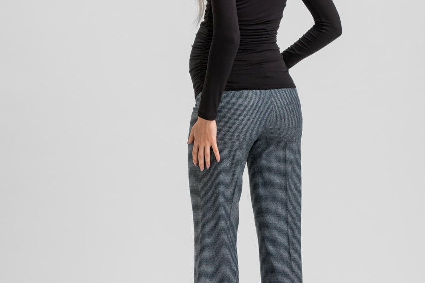 A woman shown from the back wearing a fitted black long-sleeve top, wide-leg charcoal gray trousers, and black high heels against a light gray studio background.