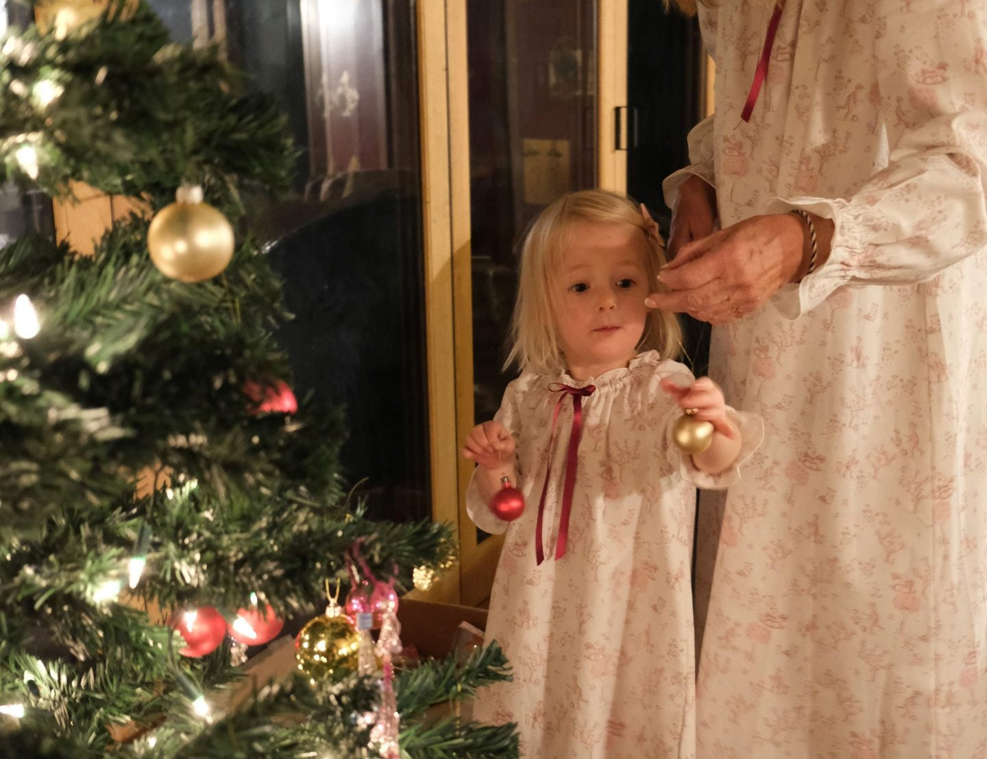 A woman and a young girl wearing matching cream nightgowns with a subtle floral print and red ribbon ties, standing barefoot by a decorated Christmas tree indoors.