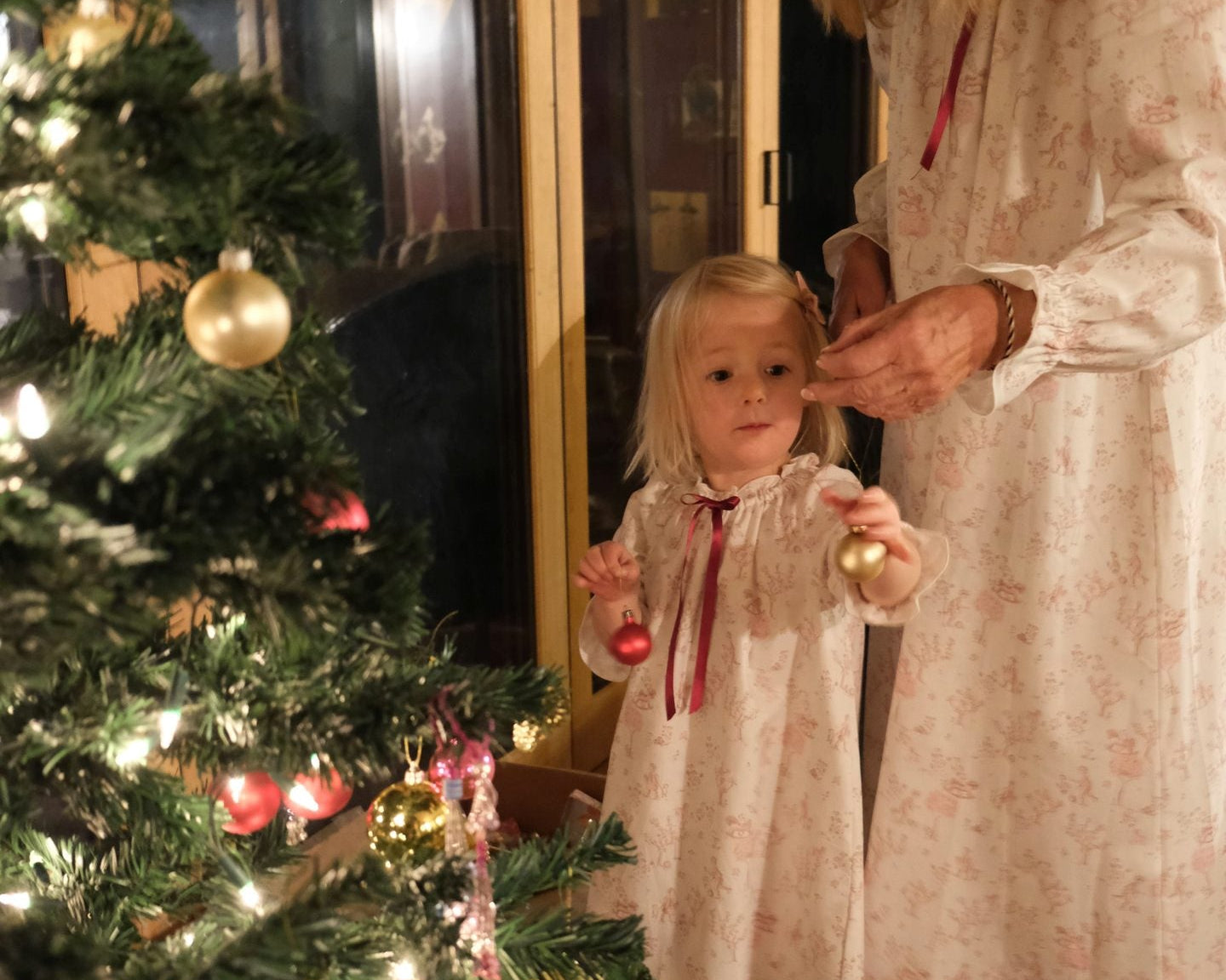 A woman and a young girl wearing matching cream nightgowns with a subtle floral print and red ribbon ties, standing barefoot by a decorated Christmas tree indoors.