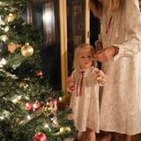 A woman and a young girl wearing matching cream nightgowns with a subtle floral print and red ribbon ties, standing barefoot by a decorated Christmas tree indoors.