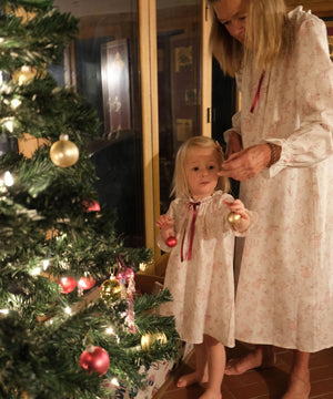 A woman and a young girl wearing matching cream nightgowns with a subtle floral print and red ribbon ties, standing barefoot by a decorated Christmas tree indoors.