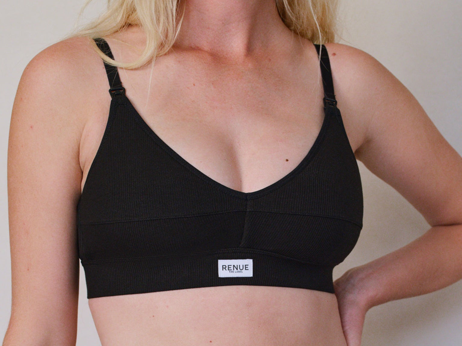 A woman wearing a black underwire bra with thin adjustable shoulder straps and a small white brand label on the front band. The brand name 'RENUE' is visible on the label.