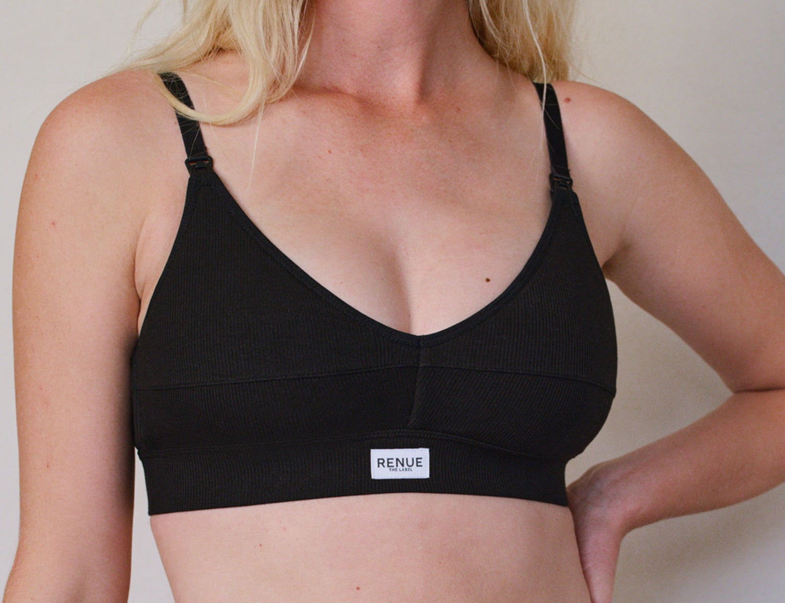 A woman wearing a black underwire bra with thin adjustable shoulder straps and a small white brand label on the front band. The brand name 'RENUE' is visible on the label.