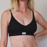 A woman wearing a black underwire bra with thin adjustable shoulder straps and a small white brand label on the front band. The brand name 'RENUE' is visible on the label.