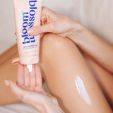 A person holding a pink tube of bloom blossom Mission Moisture Replenishing Body Cream, with white cream being dispensed onto their palm and a streak of the cream visible on their leg.