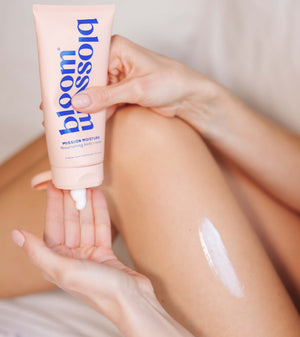 A person holding a pink tube of bloom blossom Mission Moisture Replenishing Body Cream, with white cream being dispensed onto their palm and a streak of the cream visible on their leg.
