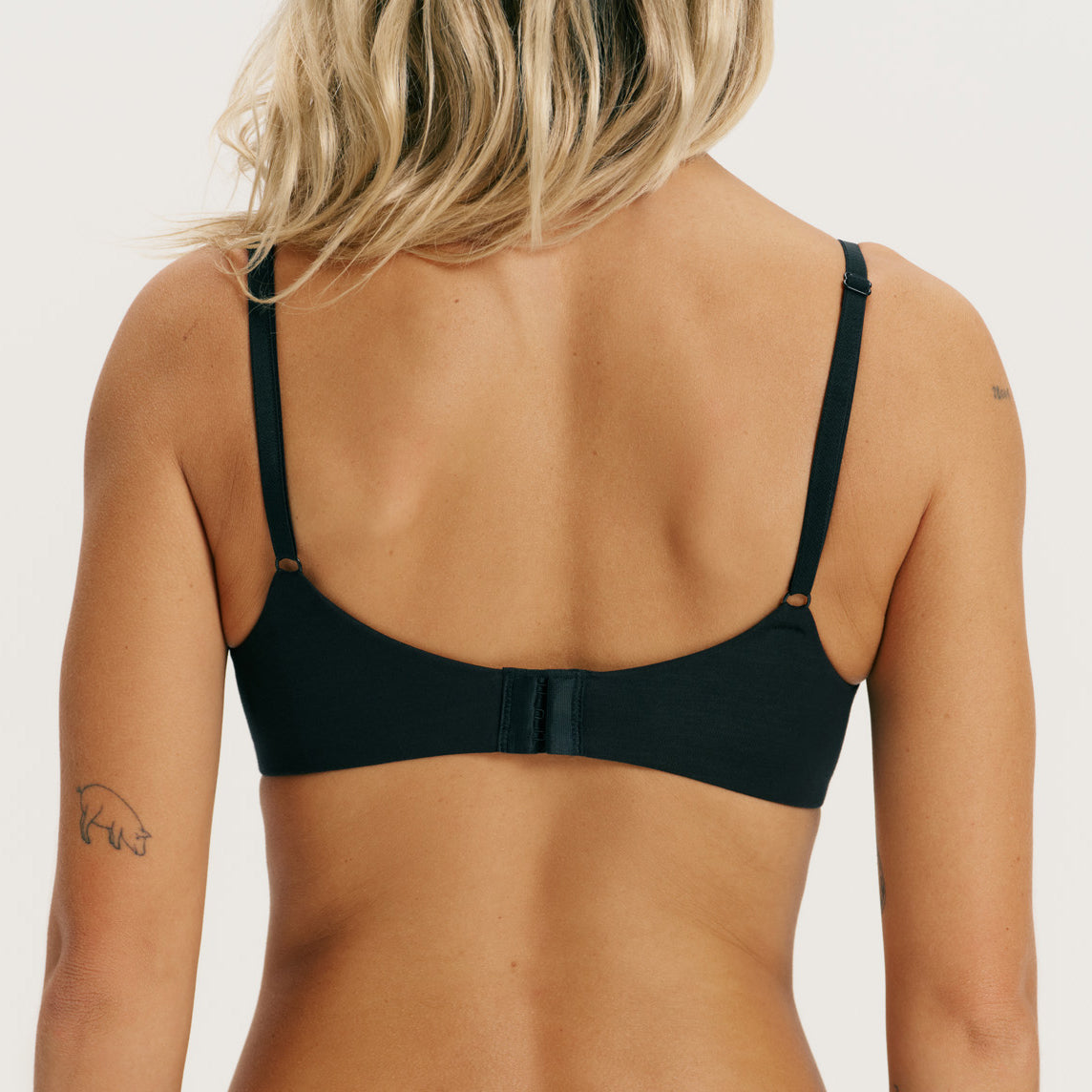 Rear view of a woman wearing a black bralette with thin adjustable straps and a center strap detail, shown against a plain light background.